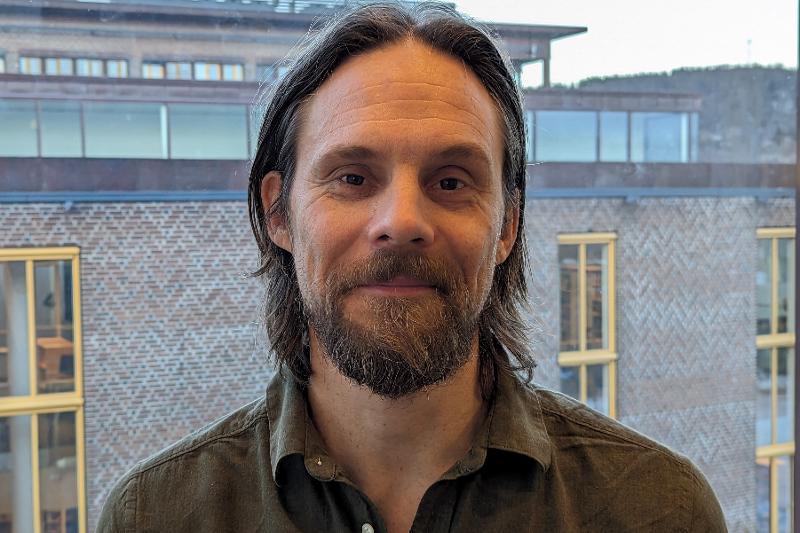 Ein mann med skjegg og halvlangt h&aring;r poserar foran eit vindauge. Ein brun mursteinsbygning i bakgrunnen.